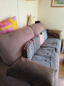 Beige sofa with a row of colorful and gray quilted cushions, brown throw on the arm, against a light wall with a framed painting above.