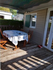 Outdoor wooden deck with a table covered by a blue-patterned tablecloth and four folding chairs, red slippers by a glass door, hedge in the background.