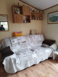 Beige patterned sofa with two colorful cushions, a quilted cover, and wooden wall shelves displaying ceramic figurines, with a framed coastal painting on the wall and wooden floor beneath.