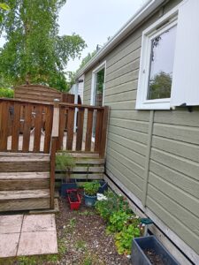 Backyard side of a house with beige siding, a wooden deck and railing, and potted plants along the wall by the windows.