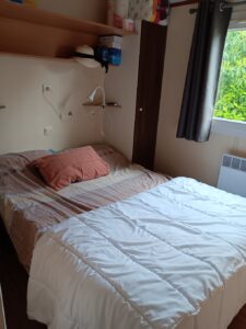 Small bedroom with a neatly made bed, white quilt, pink patterned sheets, and two pink pillows on a wooden floor.
