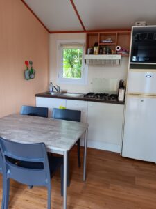 Compact caravan kitchen with a dining table and four blue chairs, sink and gas stove along the counter, fridge and microwave on the right, window above the sink with shelves nearby.