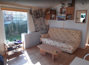 Sunlit cozy living area with a beige patterned sofa, wooden coffee table, wall shelves, and a large window showing a garden; a shoe rack sits by the door.