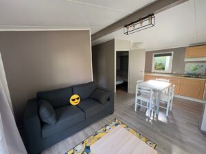 Open-plan living area with a dark gray sofa and a yellow sunglasses emoji pillow, a white dining table with chairs, and a kitchen along the right wall.
