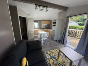 Open-plan living area with a grey sofa, white coffee table, and small dining table next to a compact kitchen with light wood cabinets and a fridge.