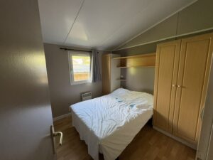 Small bedroom with a double bed, white sheets, and a wooden wardrobe on the right; shelf above the bed and a window with a curtain on the left side.