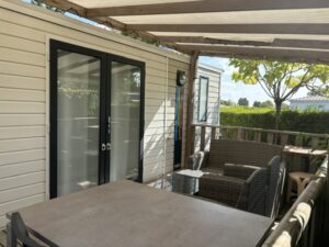 Patio seating area outside a house with double glass doors, beige siding, and a wicker sofa set under a wooden pergola. A glass-topped table sits in front.