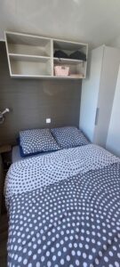 Small bedroom with a double bed in polka dot blue and white bedding, two pillows, shelves above the headboard, and a white wardrobe nearby.