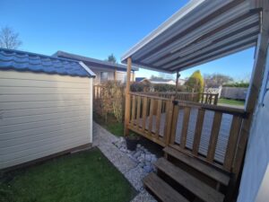 Backyard with a wooden deck and stairs under a metal canopy beside a beige shed; gravel path, potted plant, and lawn visible under a blue sky.