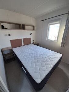 Bedroom with a double bed featuring two brown headboard panels, matching nightstands, and a wall shelf above the bed. A window with curtains is on the right.