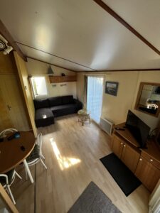 Cozy living room in a caravan with a black sectional sofa, wooden cabinets, and a small round glass table near a window with sheer curtains in natural light