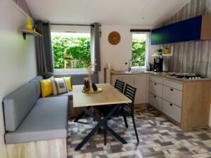 Cozy kitchen-dining nook with grey sofa, yellow cushions, and a wooden table facing a window with greenery outside