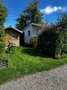 Small wooden shed beside a cream house with a white window, a bicycle leaning against the shed, green lawn and shrubs, and a gravel path in the foreground under a blue sky.