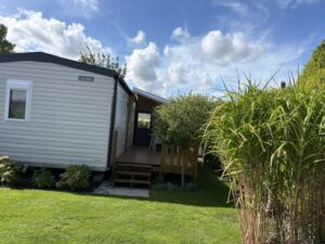 White mobile home with a wooden deck and steps, set on a green lawn under a blue, cloudy sky.
